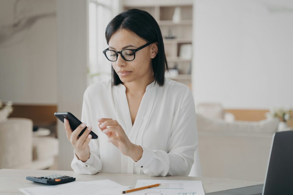 Businesswoman in glasses using mobile phone apps for business time management sitting at laptop